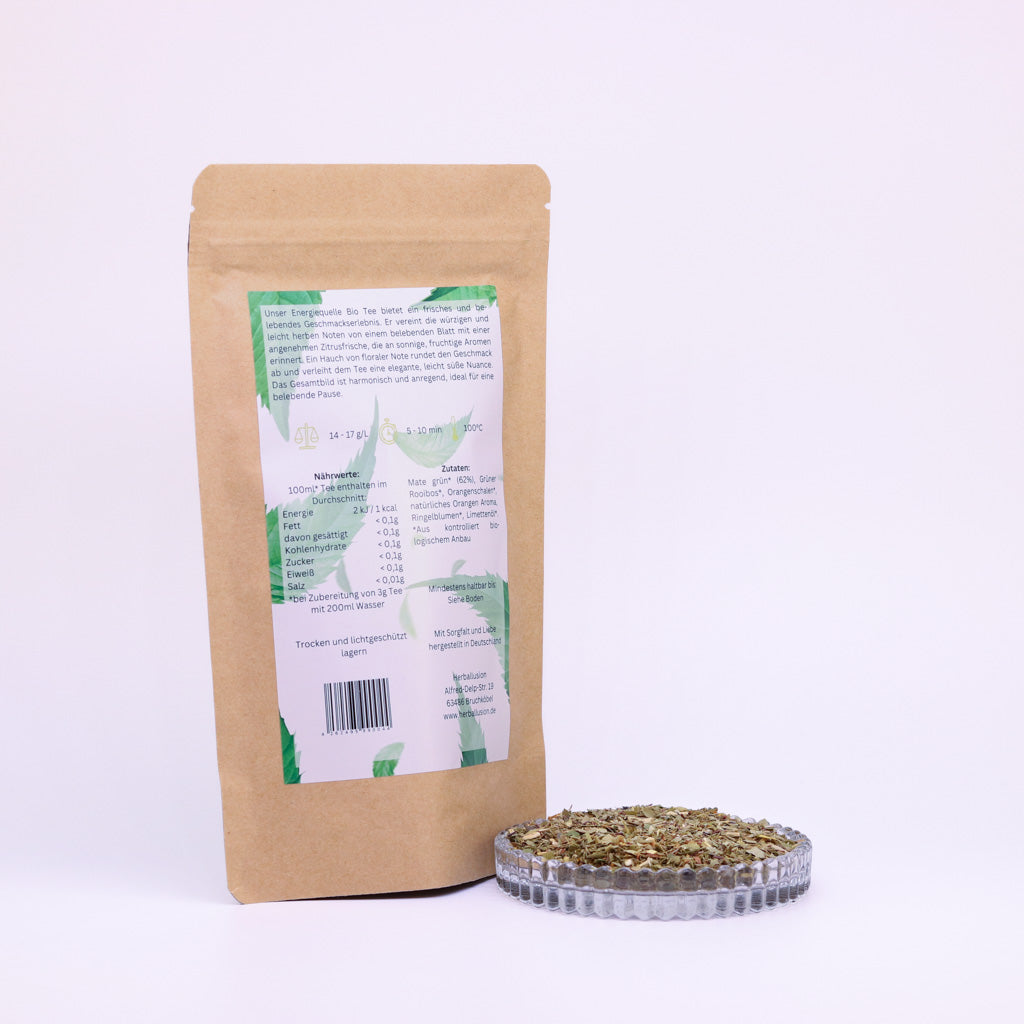 100g-Beutel Bio-Matetee ‚Energiequelle‘ mit gelb-grünem Etikett Rückseite, daneben Glasschale mit losem Tee.
