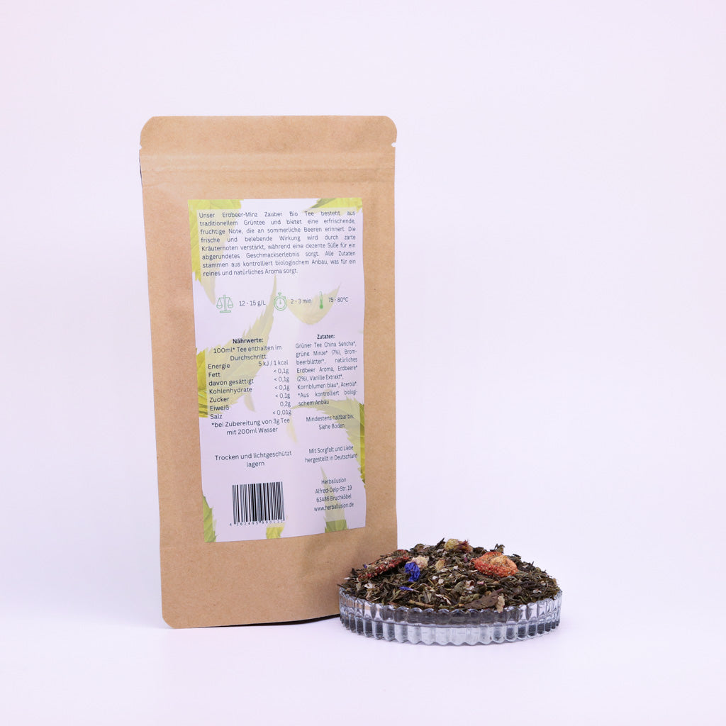 100g-Beutel Bio-Grüntee ‚Erdbeer-Minz Zauber‘ mit gelb-grünem Etikett Rückseite, daneben Glasschale mit losem Tee.