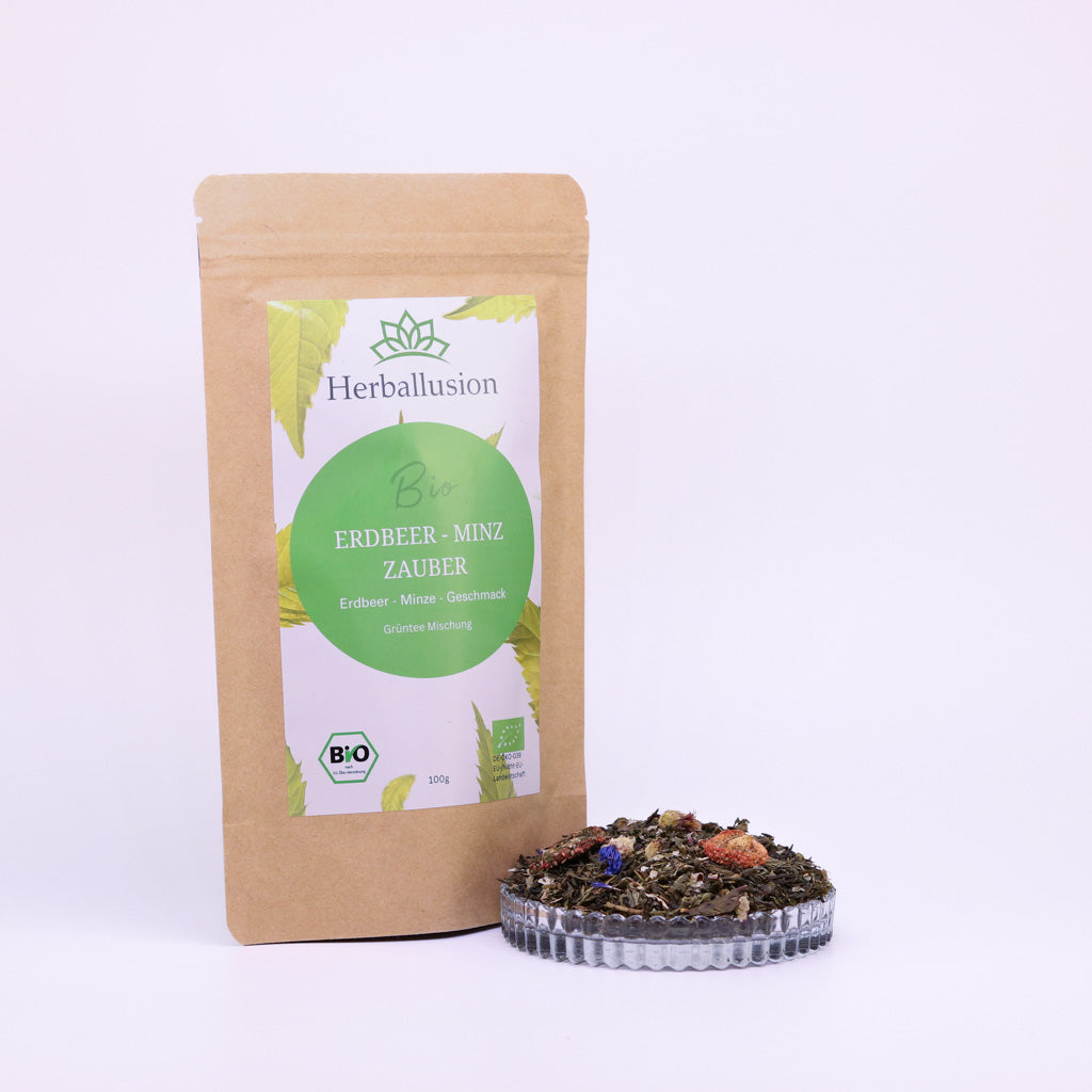 100g-Beutel Bio-Grüntee ‚Erdbeer-Minz Zauber‘ mit gelb-grünem Etikett, daneben Glasschale mit losem Tee.