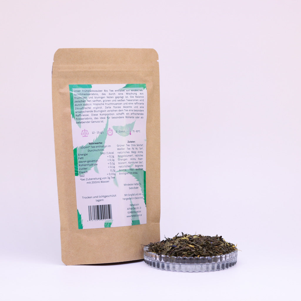 100g-Beutel Bio-Grüntee ‚Frühstückszauber‘ mit rosa-grünem Etikett Rückseite, daneben Glasschale mit losem Tee.
