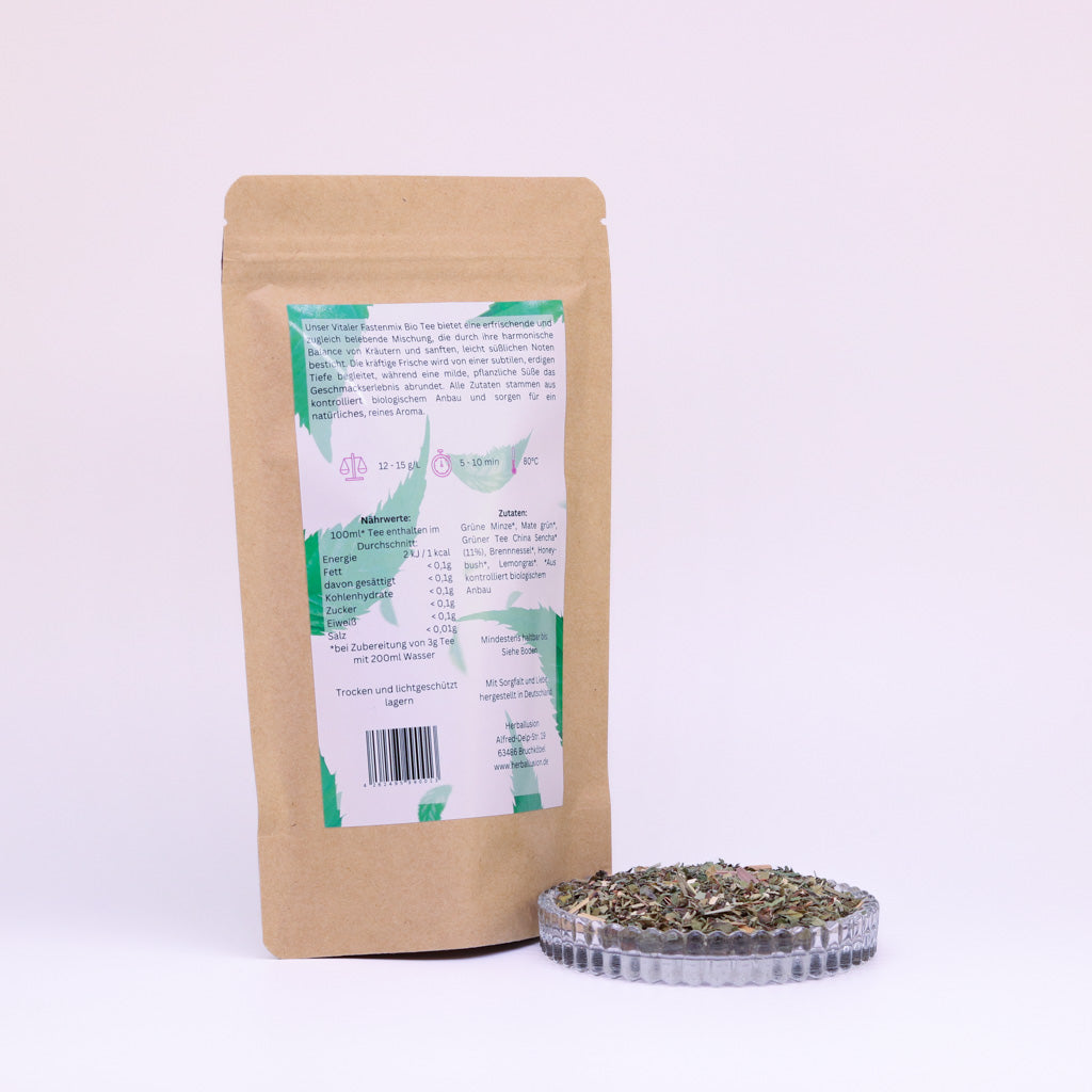 100g-Beutel Bio-Grüntee ‚Vitaler Fastenmix‘ mit rosa-grünem Etikett Rückseite, daneben Glasschale mit losem Tee.