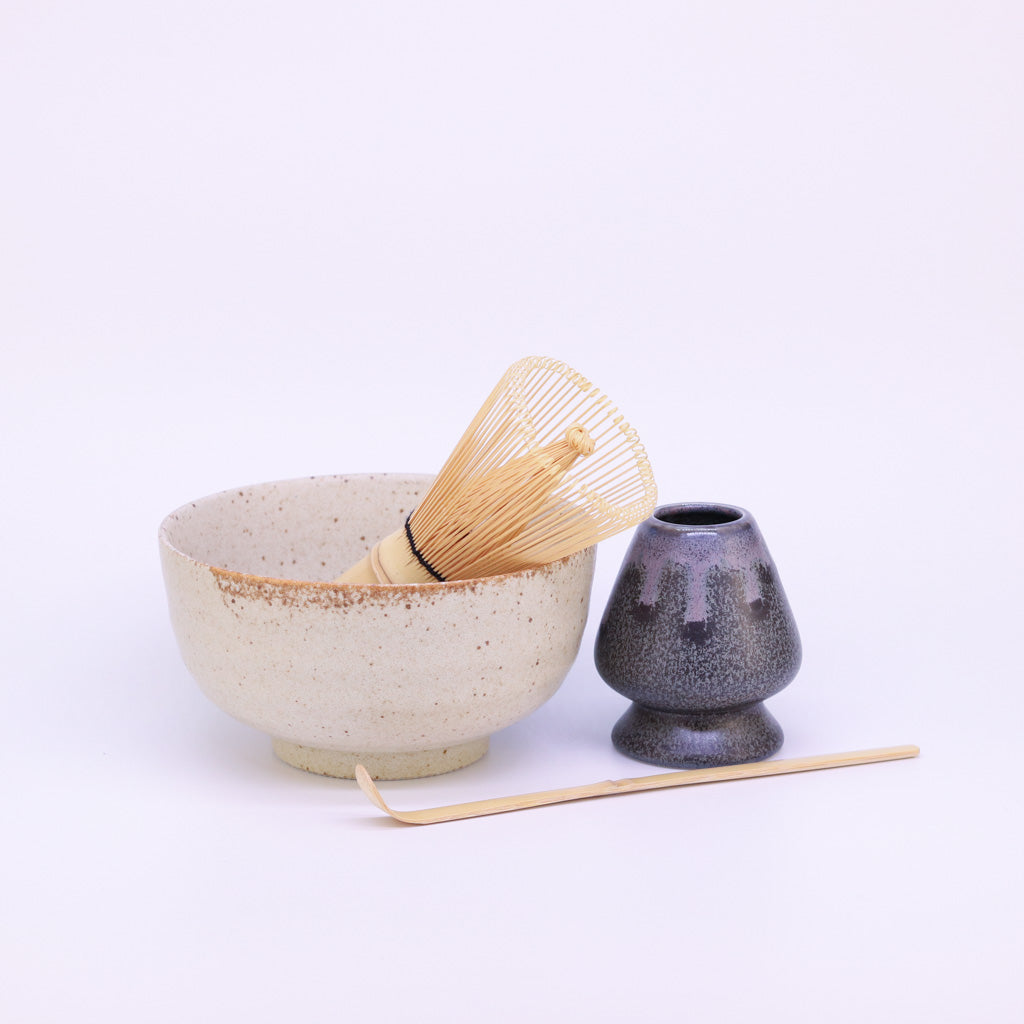 Matcha-Set auf weißem Hintergrund: Eine cremefarbene Matcha-Schale mit einem Matcha-Besen, dessen Borsten aus der Schale herausragen. Neben der Schale steht der Matcha-Besenhalter, und davor liegt der vollständig sichtbare Bambus-Löffel, längs platziert