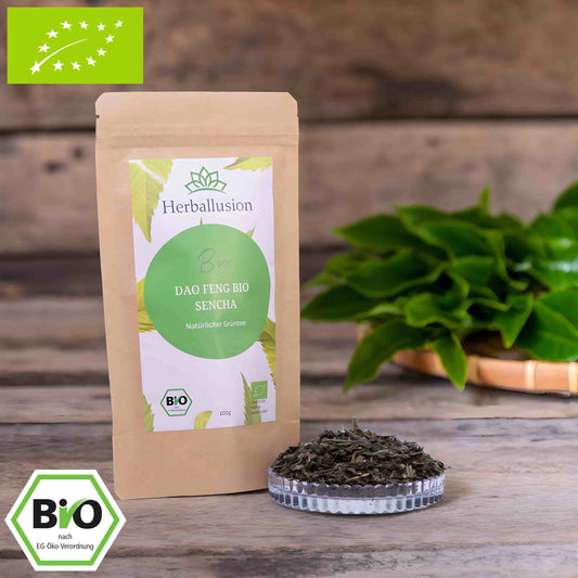 100g-Beutel Bio-Grüntee ‚Dao Feng Bio Sencha‘ mit gelb-grünem Etikett, daneben Glasschale mit losem Tee. Im Hintergrund Zutaten zu sehen sowie Bio und EU Zertifizierung.
