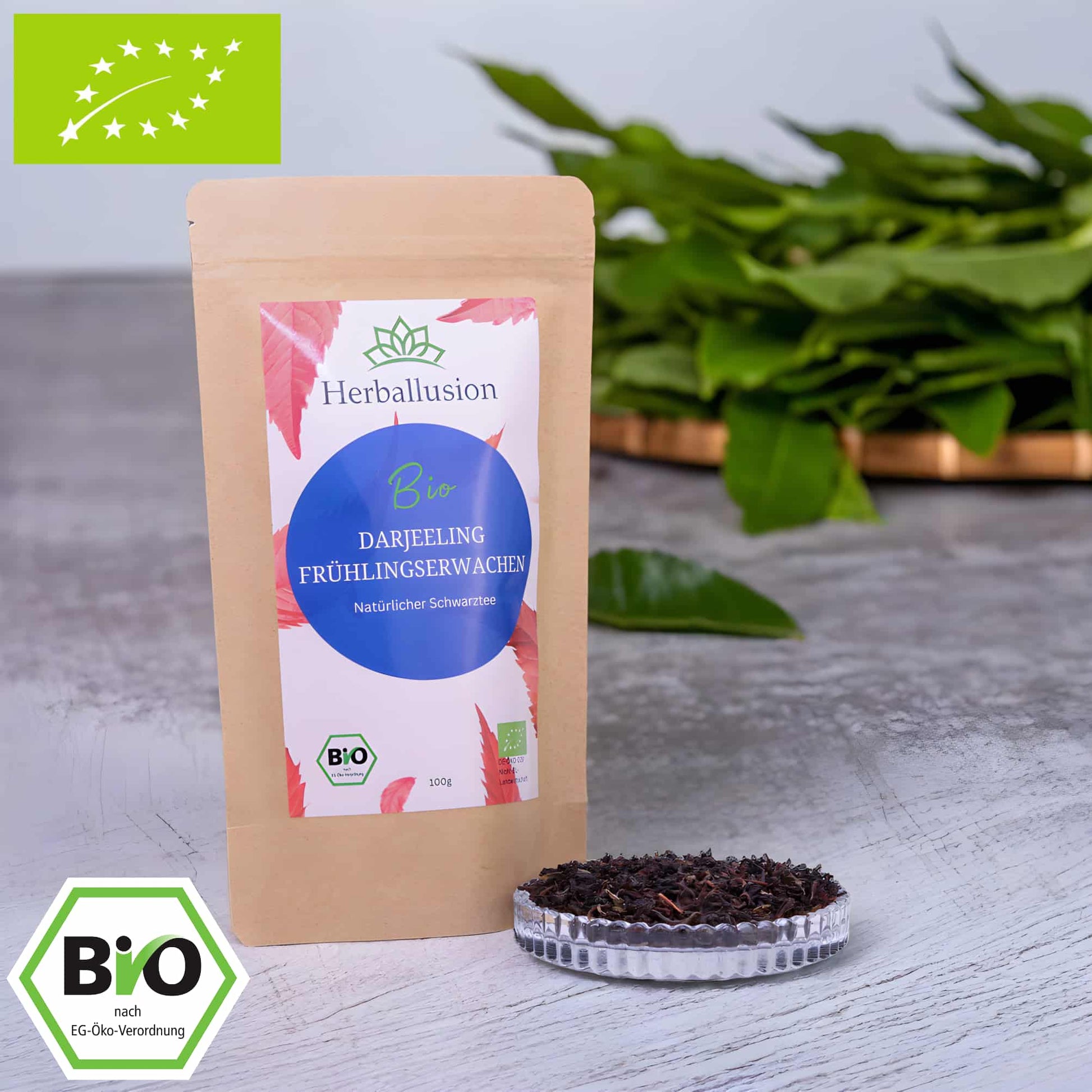 100g-Beutel Bio-Schwarztee ‚Darjeeling Frühlingserwachen‘ mit rot-blauem Etikett, daneben Glasschale mit losem Tee. Im Hintergrund Zutaten zu sehen sowie Bio und EU Zertifizierung.