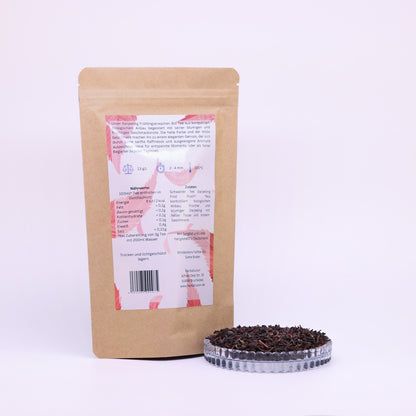 100g-Beutel Bio-Schwarztee ‚Darjeeling Frühlingserwachen‘ mit rot-blauem Etikett Rückseite, daneben Glasschale mit losem Tee.