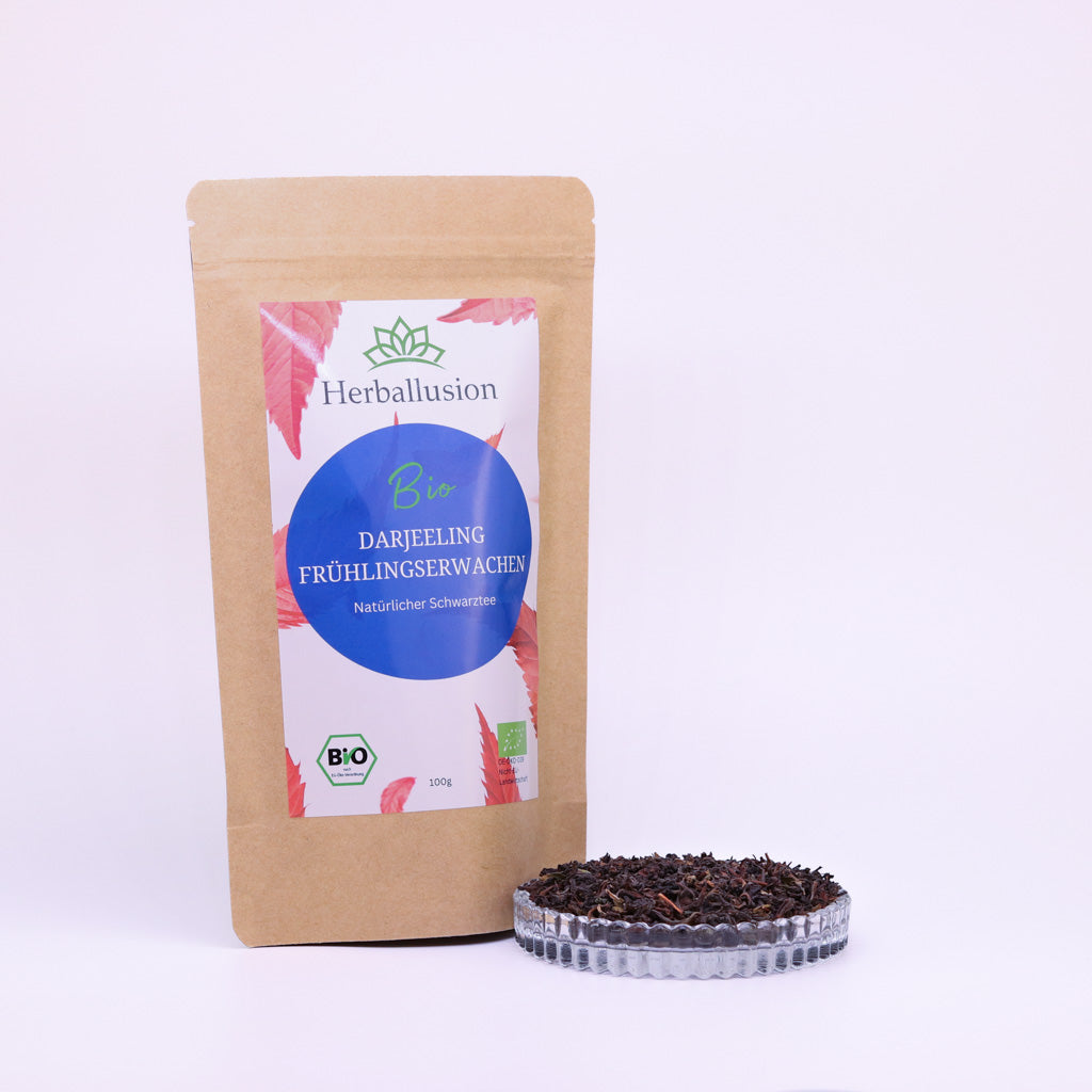 100g-Beutel Bio-Schwarztee ‚Darjeeling Frühlingserwachen‘ mit rot-blauem Etikett, daneben Glasschale mit losem Tee.