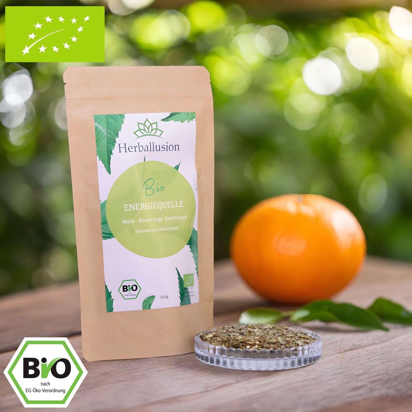 100g-Beutel Bio-Matetee ‚Energiequelle‘ mit gelb-grünem Etikett, daneben Glasschale mit losem Tee. Im Hintergrund Zutaten zu sehen sowie Bio und EU Zertifizierung.