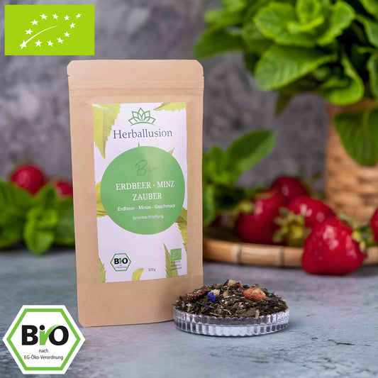 100g-Beutel Bio-Grüntee ‚Erdbeer-Minz Zauber‘ mit gelb-grünem Etikett, daneben Glasschale mit losem Tee. Im Hintergrund Zutaten zu sehen sowie Bio und EU Zertifizierung.