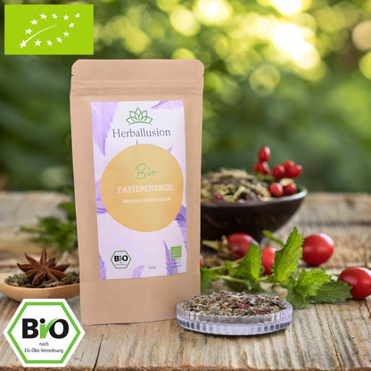 100g-Beutel Bio-Kräutertee ‚Fastenenergie‘ mit orange-lilanem Etikett, daneben Glasschale mit losem Tee. Im Hintergrund Zutaten zu sehen sowie Bio und EU Zertifizierung.