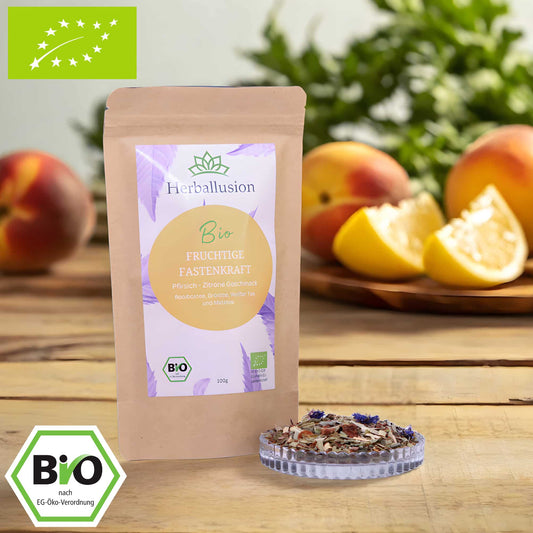 100g-Beutel Bio-Rooibostee ‚Fruchtige Fastenkraft‘ mit orange-lilanem Etikett, daneben Glasschale mit losem Tee. Im Hintergrund Zutaten zu sehen sowie Bio und EU Zertifizierung.