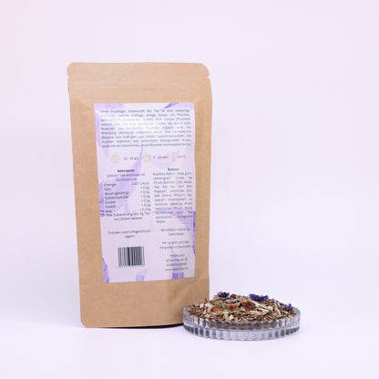 100g-Beutel Bio-Rooibostee ‚Fruchtige Fastenkraft‘ mit orange-lilanem Etikett Rückseite, daneben Glasschale mit losem Tee.