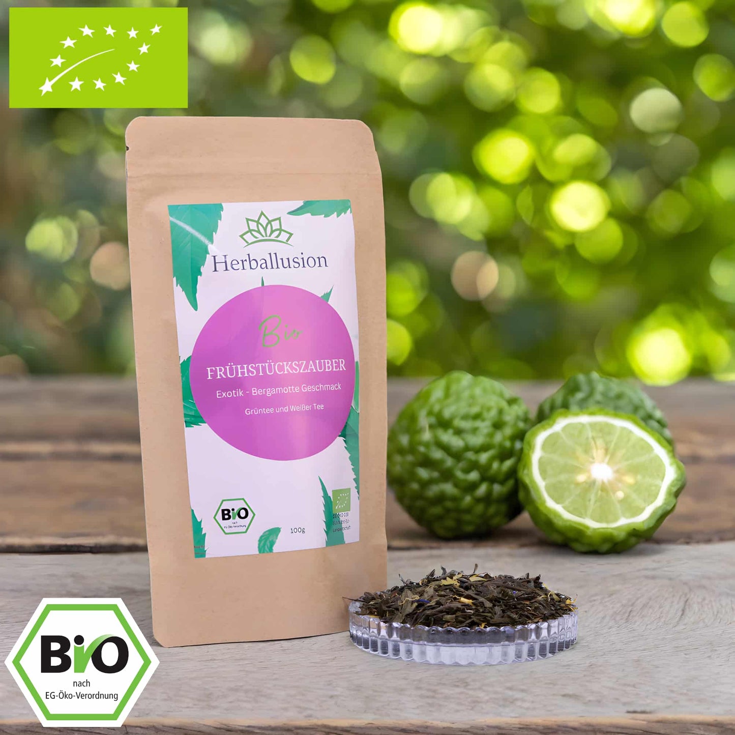 100g-Beutel Bio-Grüntee ‚Frühstückszauber‘ mit rosa-grünem Etikett, daneben Glasschale mit losem Tee. Im Hintergrund Zutaten zu sehen sowie Bio und EU Zertifizierung.