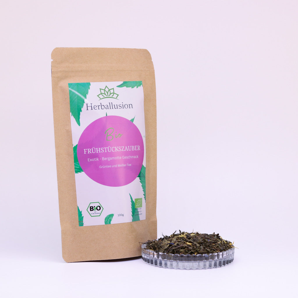 100g-Beutel Bio-Grüntee ‚Frühstückszauber‘ mit rosa-grünem Etikett, daneben Glasschale mit losem Tee.