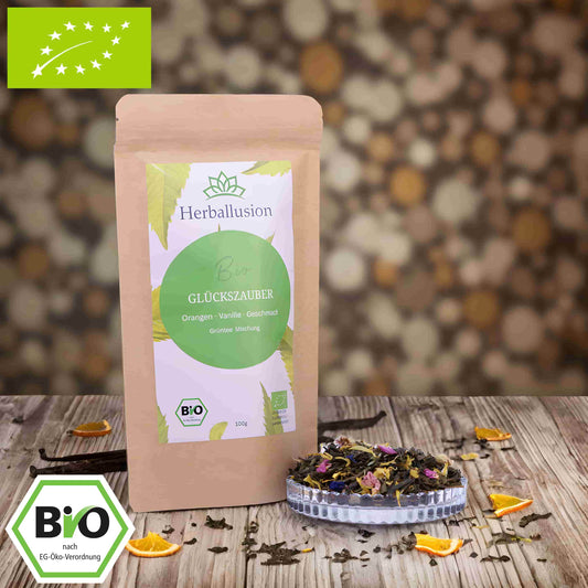 100g-Beutel Bio-Grüntee ‚Glückszauber‘ mit gelb-grünem Etikett, daneben Glasschale mit losem Tee. Im Hintergrund Zutaten zu sehen sowie Bio und EU Zertifizierung.