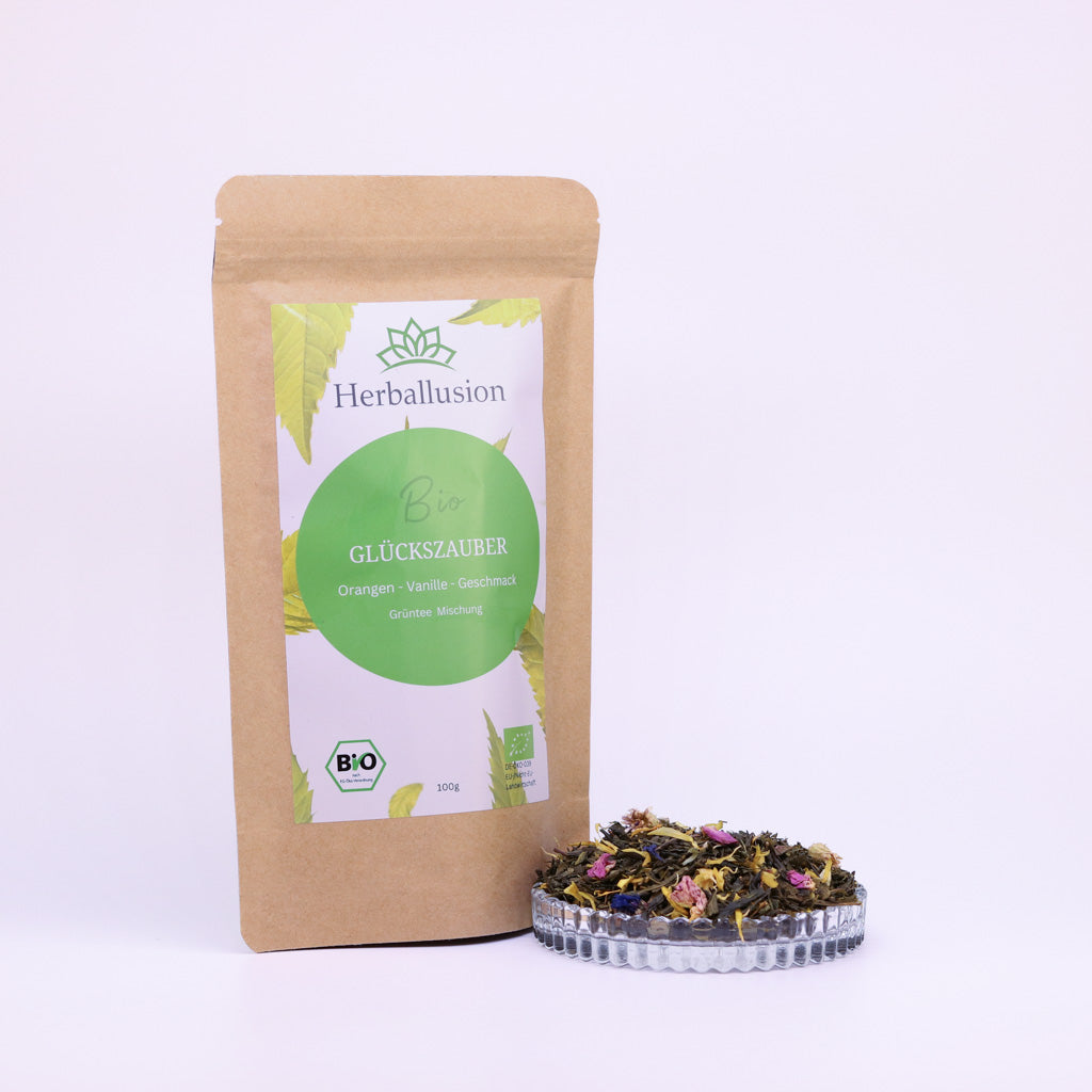 100g-Beutel Bio-Grüntee ‚Glückszauber‘ mit gelb-grünem Etikett, daneben Glasschale mit losem Tee.