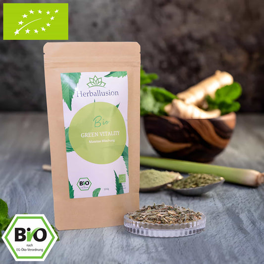 100g-Beutel Bio-Matetee ‚Green Vitality‘ mit gelb-grünem Etikett, daneben Glasschale mit losem Tee. Im Hintergrund Zutaten zu sehen sowie Bio und EU Zertifizierung.