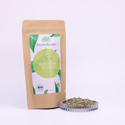 100g-Beutel Bio-Matetee ‚Mate-Elixier‘ mit gelb-grünem Etikett, daneben Glasschale mit losem Tee.