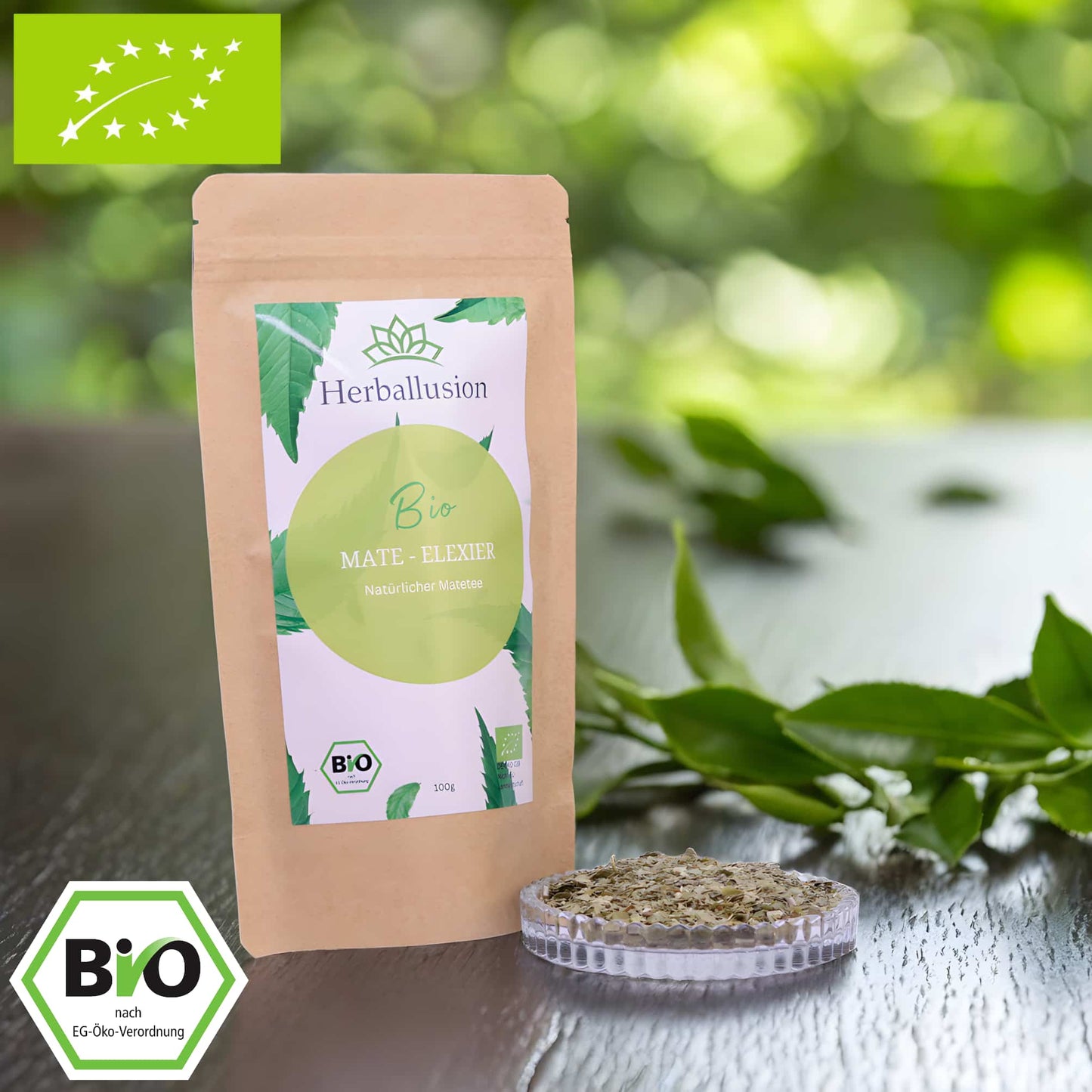 100g-Beutel Bio-Matetee ‚Mate-Elixier‘ mit gelb-grünem Etikett, daneben Glasschale mit losem Tee. Im Hintergrund Zutaten zu sehen sowie Bio und EU Zertifizierung.