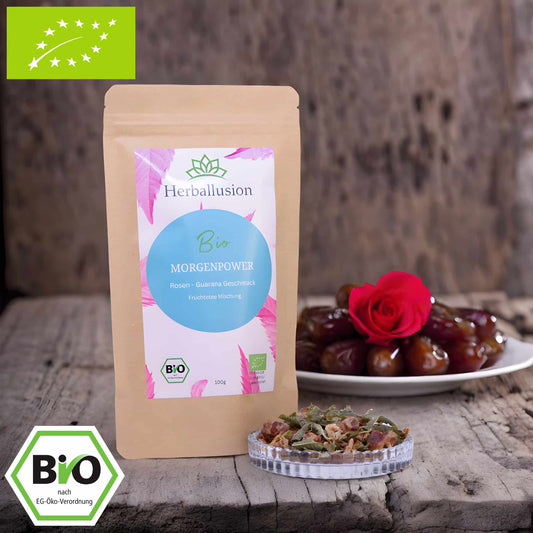 100g-Beutel Bio-Früchtetee ‚Morgenpower‘ mit rosa-blauem Etikett, daneben Glasschale mit losem Tee. Im Hintergrund Zutaten zu sehen sowie Bio und EU Zertifizierung.