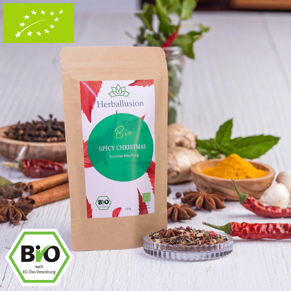 100g-Beutel Bio-Grüntees‚Spicy Christmas‘ mit rot-grünem Etikett, daneben Glasschale mit losem Tee.Im Hintergrund Zutaten zu sehen sowie Bio und EU Zertifizierung.