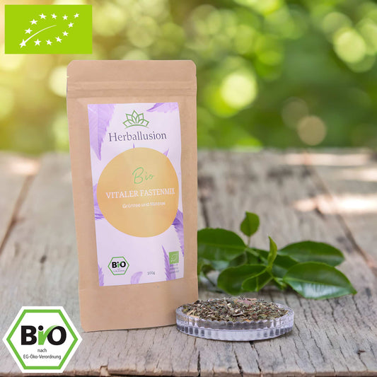 100g-Beutel Bio-Grüntee ‚Vitaler Fastenmix‘ mit orange-lilanem Etikett, daneben Glasschale mit losem Tee. Im Hintergrund Zutaten zu sehen sowie Bio und EU Zertifizierung.