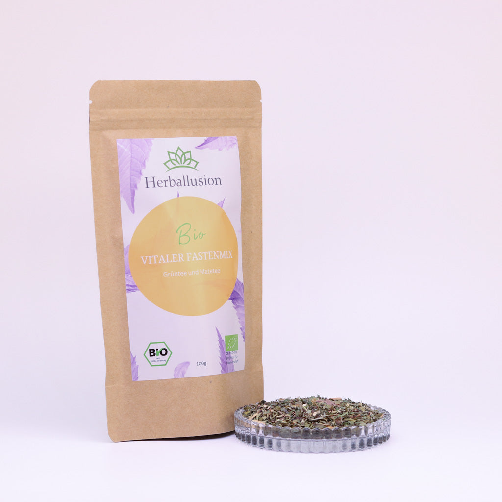 100g-Beutel Bio-Grüntee ‚Vitaler Fastenmix‘ mit orange-lilanem Etikett, daneben Glasschale mit losem Tee.