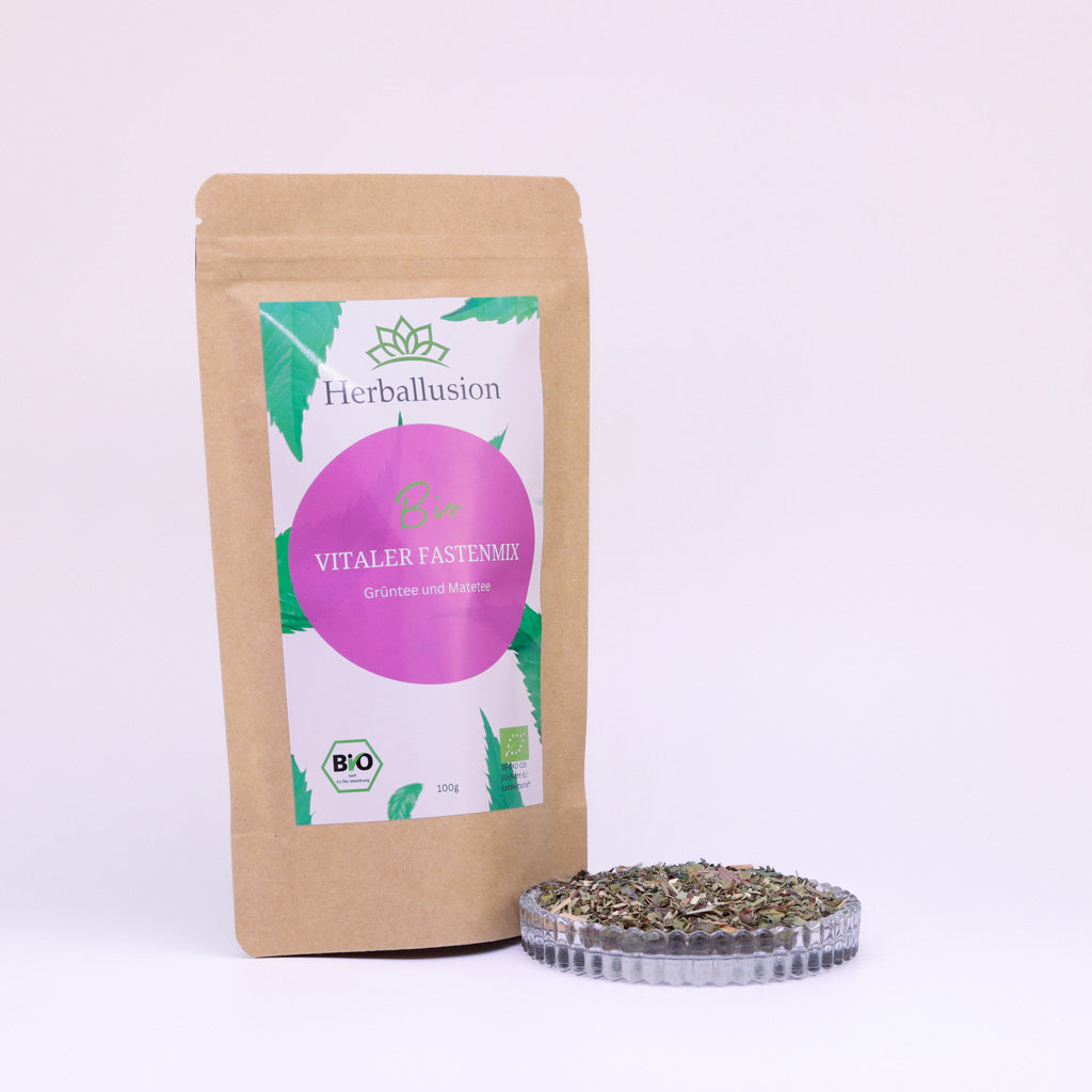 100g-Beutel Bio-Grüntee ‚Vitaler Fastenmix‘ mit rosa-grünem Etikett, daneben Glasschale mit losem Tee.