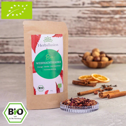 100g-Beutel Bio-Früchtetee ‚Weihnachtszauber‘ mit rot-grünem Etikett, daneben Glasschale mit losem Tee.  Im Hintergrund Zutaten zu sehen sowie Bio und EU Zertifizierung.
