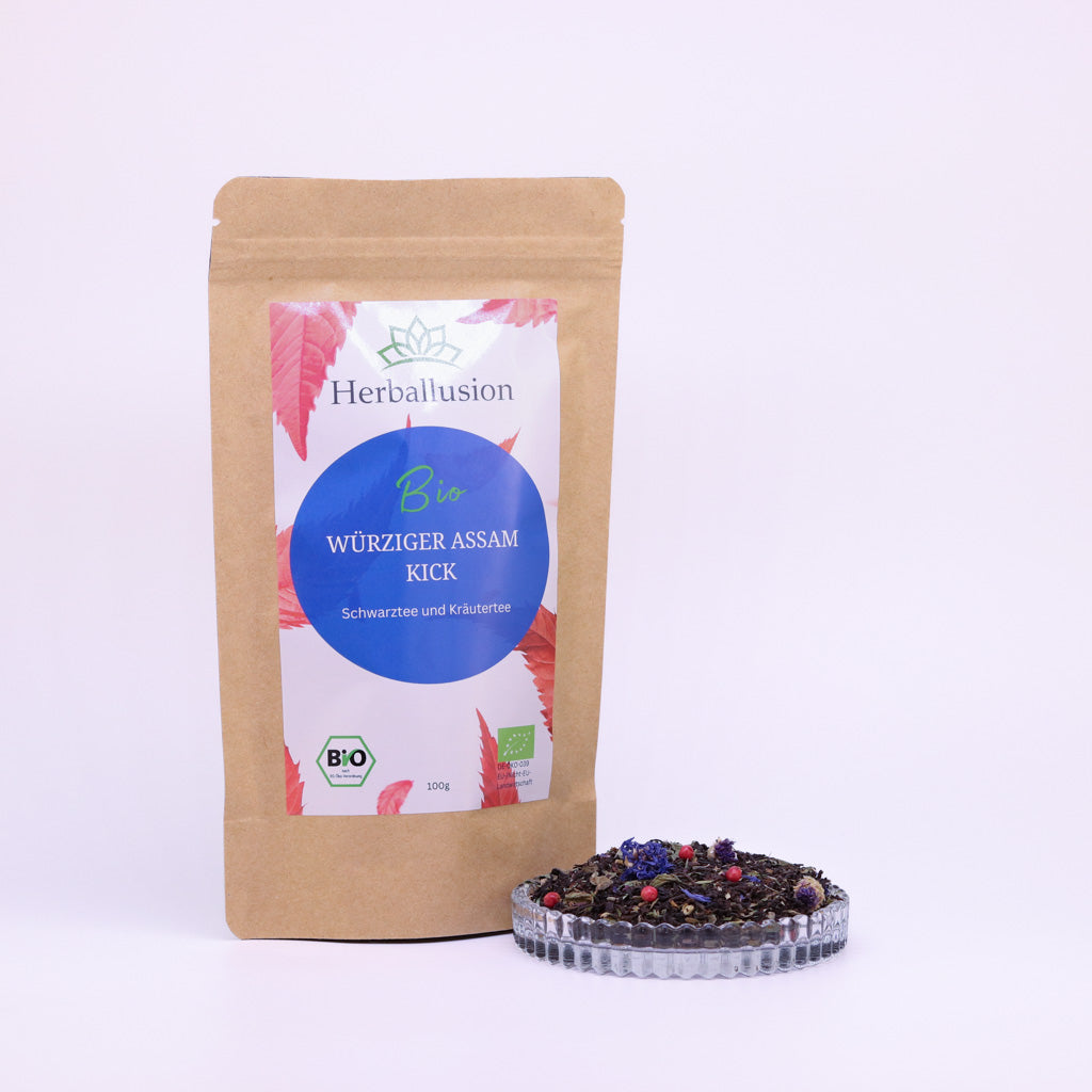100g-Beutel Bio-Schwarztee ‚Würziger Assam Kick‘ mit rot-blauem Etikett, daneben Glasschale mit losem Tee.