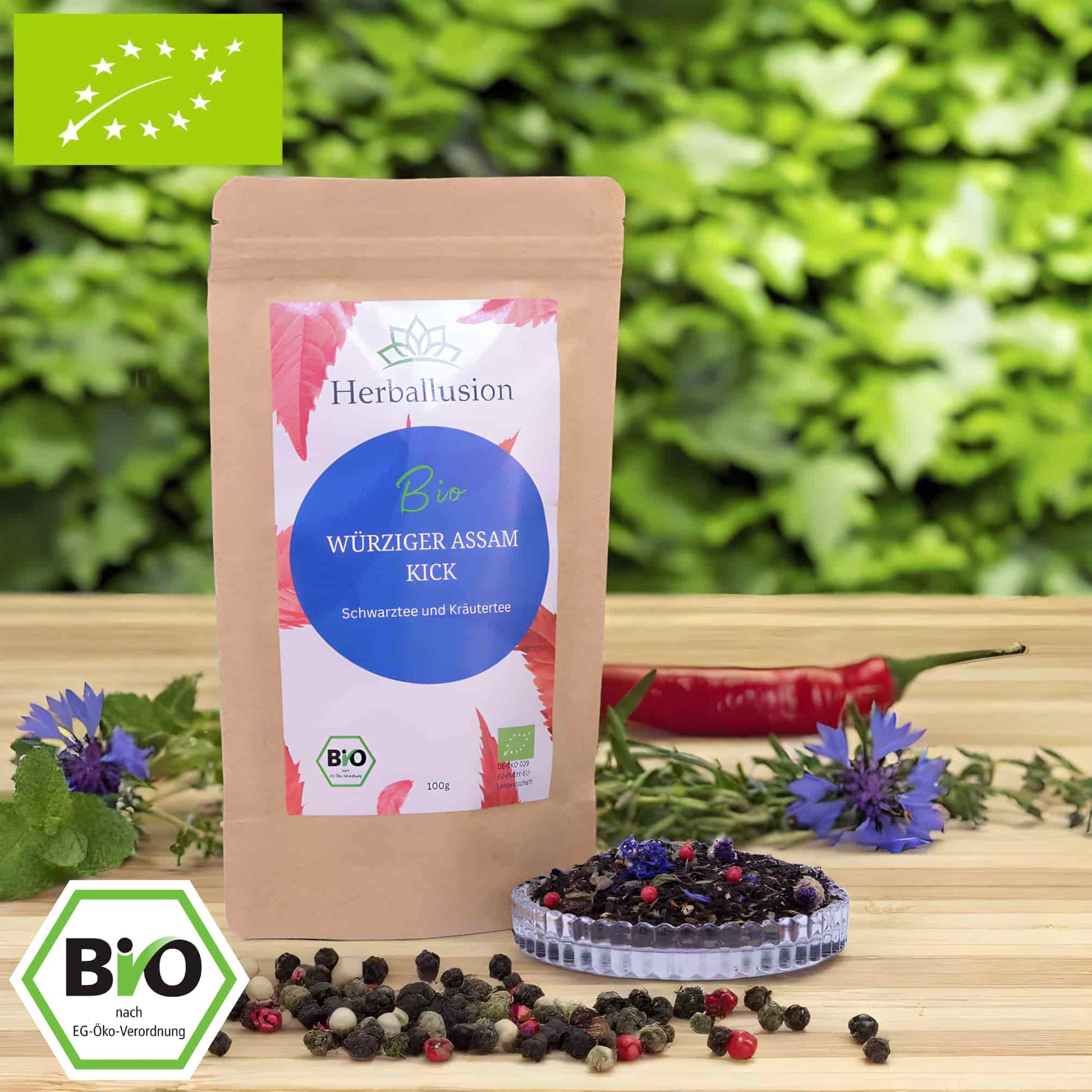 100g-Beutel Bio-Schwarztee ‚Würziger Assam Kick‘ mit rot-blauem Etikett, daneben Glasschale mit losem Tee. Im Hintergrund Zutaten zu sehen sowie Bio und EU Zertifizierung.