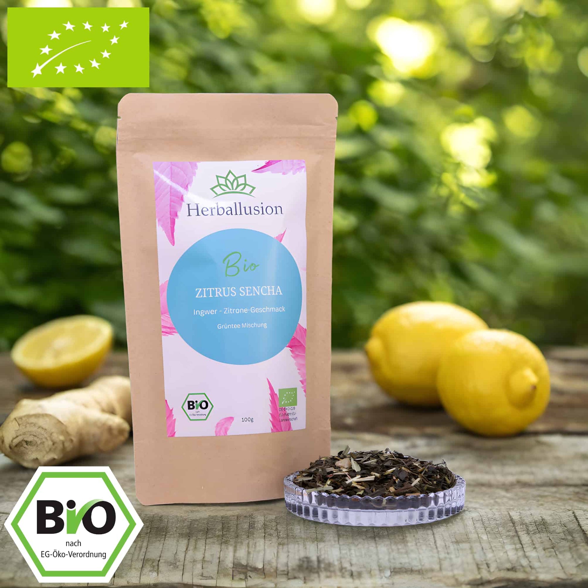 100g-Beutel Bio-Grüntee ‚Zitrus Sencha‘ mit rosa-blauem Etikett, daneben Glasschale mit losem Tee. Im Hintergrund Zutaten zu sehen sowie Bio und EU Zertifizierung.