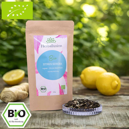 100g-Beutel Bio-Grüntee ‚Zitrus Sencha‘ mit rosa-blauem Etikett, daneben Glasschale mit losem Tee. Im Hintergrund Zutaten zu sehen sowie Bio und EU Zertifizierung.