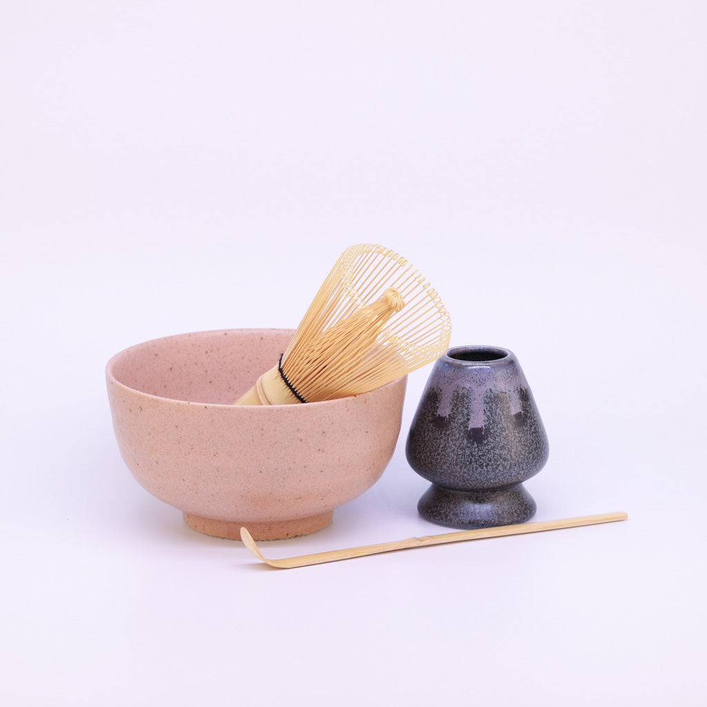 Matcha-Set auf weißem Hintergrund: Eine orangefarbene Matcha-Schale mit einem Matcha-Besen, dessen Borsten aus der Schale herausragen. Neben der Schale steht der Matcha-Besenhalter, und davor liegt der vollständig sichtbare Bambus-Löffel, längs platziert