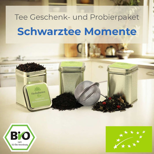 Drei nachhaltige Mini-Teedosen mit losem Bio-Schwarztee stehen auf einer Küchentheke; eine Dose ist geöffnet und zeigt den Tee, vor den anderen liegt der lose Tee zusammen mit einem Edelstahlsieb – im Hintergrund ist eine moderne Küche zu sehen. 