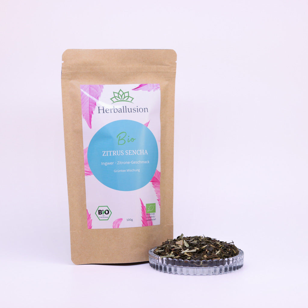 100g-Beutel Bio-Grüntee ‚Zitrus Sencha‘ mit rosa-blauem Etikett, daneben Glasschale mit losem Tee.